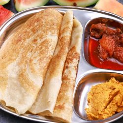 3 dosas made with watermelon skin and rice served in a steel plate with tomato chutney and pickle. Watermelon pieces placed around the plate for decoration.